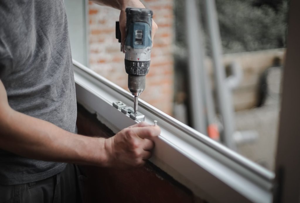 Un homme caucasien installe des ferrures pour un cadre de fenêtre à l'aide d'une perceuse.