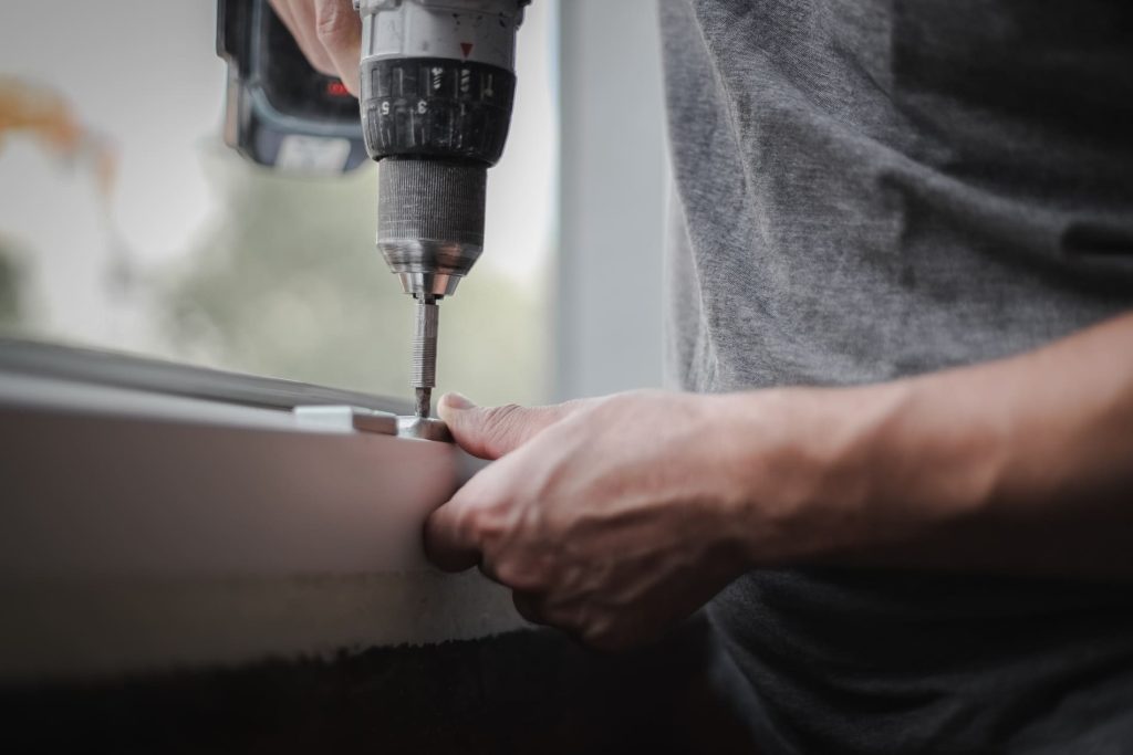 Un jeune homme caucasien installe des ferrures métalliques sur un cadre de fenêtre en PVC à l'aide d'une perceuse, serrant une vis dans une pièce en travaux.