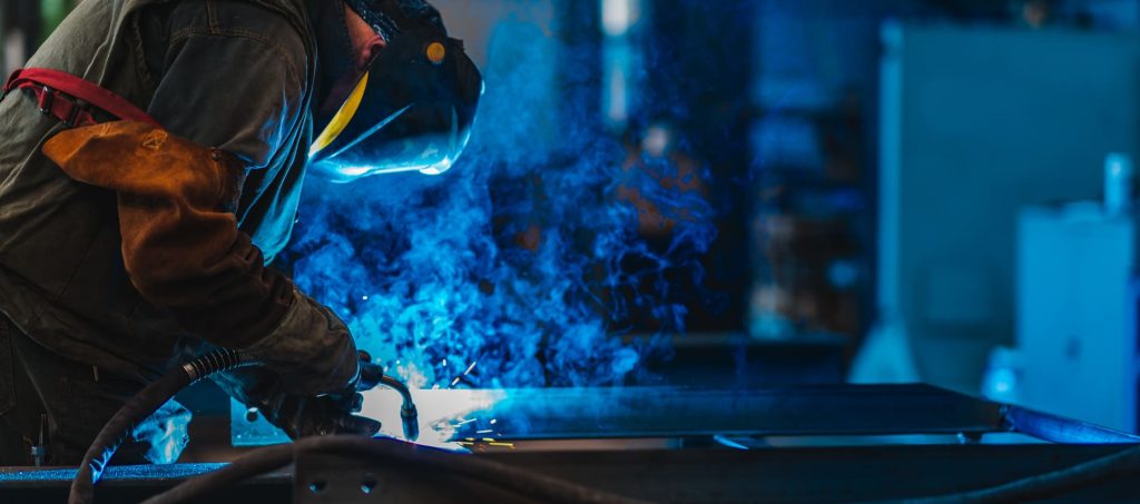 Soudeur industriel avec torche et casque de protection dans un grand hall, soudage de profilés métalliques